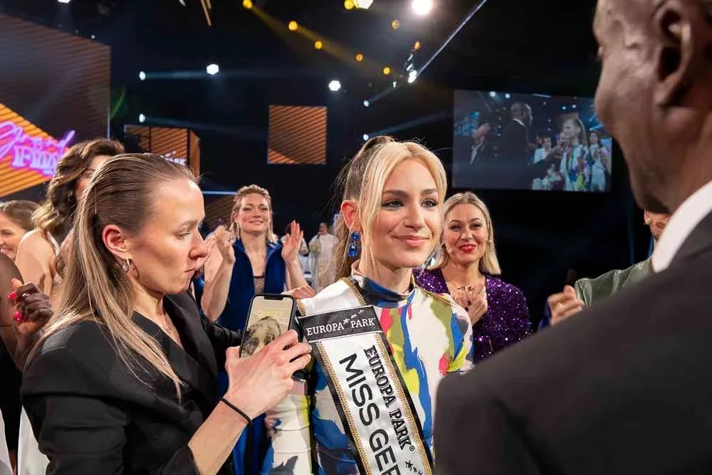 Die Siegerin inmitten applaudierender Finalistinnen, im Hintergrund die Bühnenleinwand