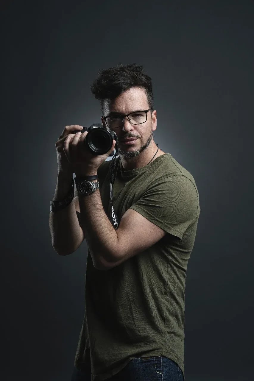 Porträt vor dunklem Hintergrund: ein Mann mit Brille und olivgrünem T-Shirt hält eine Sony-Kamera vor das Gesicht und blickt am Objektiv vorbei in die Kamera