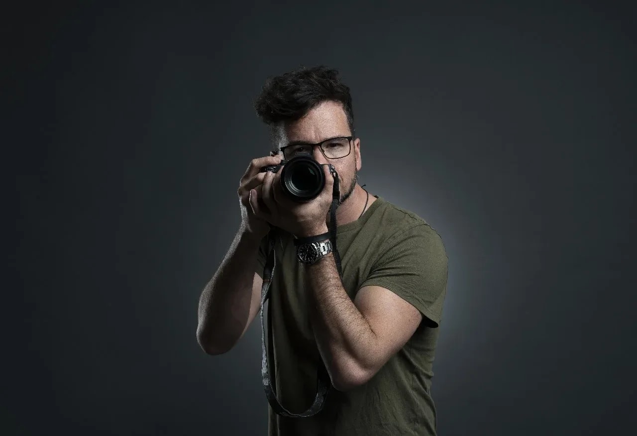 Ein Mann mit Brille und olivgrünem T-Shirt richtet eine Kamera direkt auf den Betrachter, dunkler Studiohintergrund